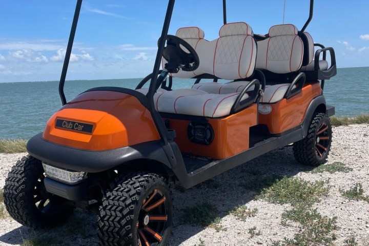 Orange golf cart with black tires on a beachside path under a blue sky.