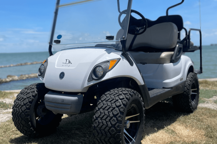 White golf cart with large tires on grass near water and blue sky.